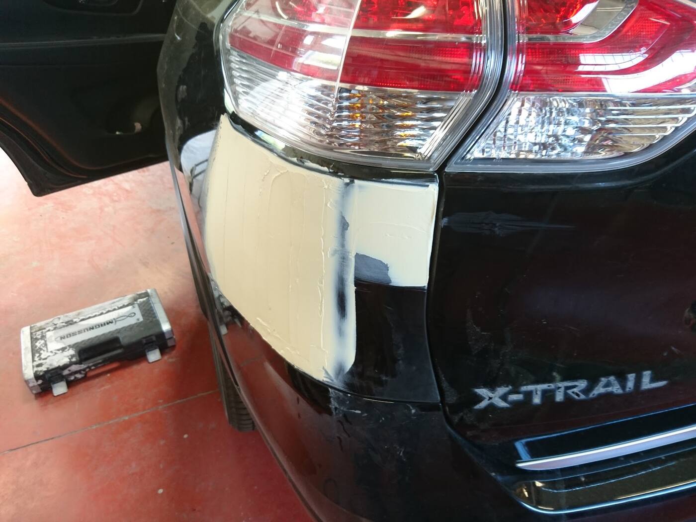 Aile arrière du X-Trail reprise au mastic beige, juste sous le feu, sur le sol rouge de l'atelier